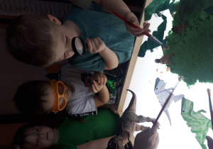 Na zdjęciu widać przedszkolaki bawiące się w paleontologów, chłopiec trzyma lupę, zabawa na stoliku podświetlanym